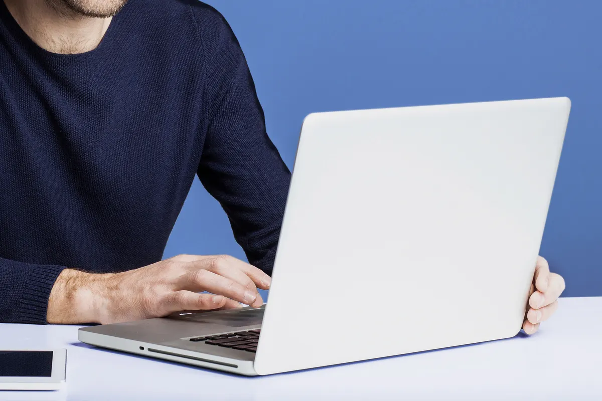 Homem de suéter azul-marinho digitando em um notebook sobre uma mesa branca